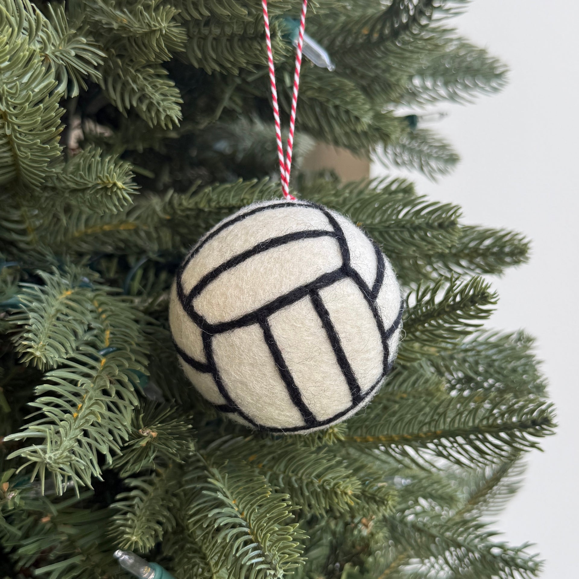 A felt Christmas ornament shaped like a volleyball hangs on a green evergreen tree. The ornament is cream-colored with black stitched panel lines and is suspended by a red-and-white striped string against a light gray background.