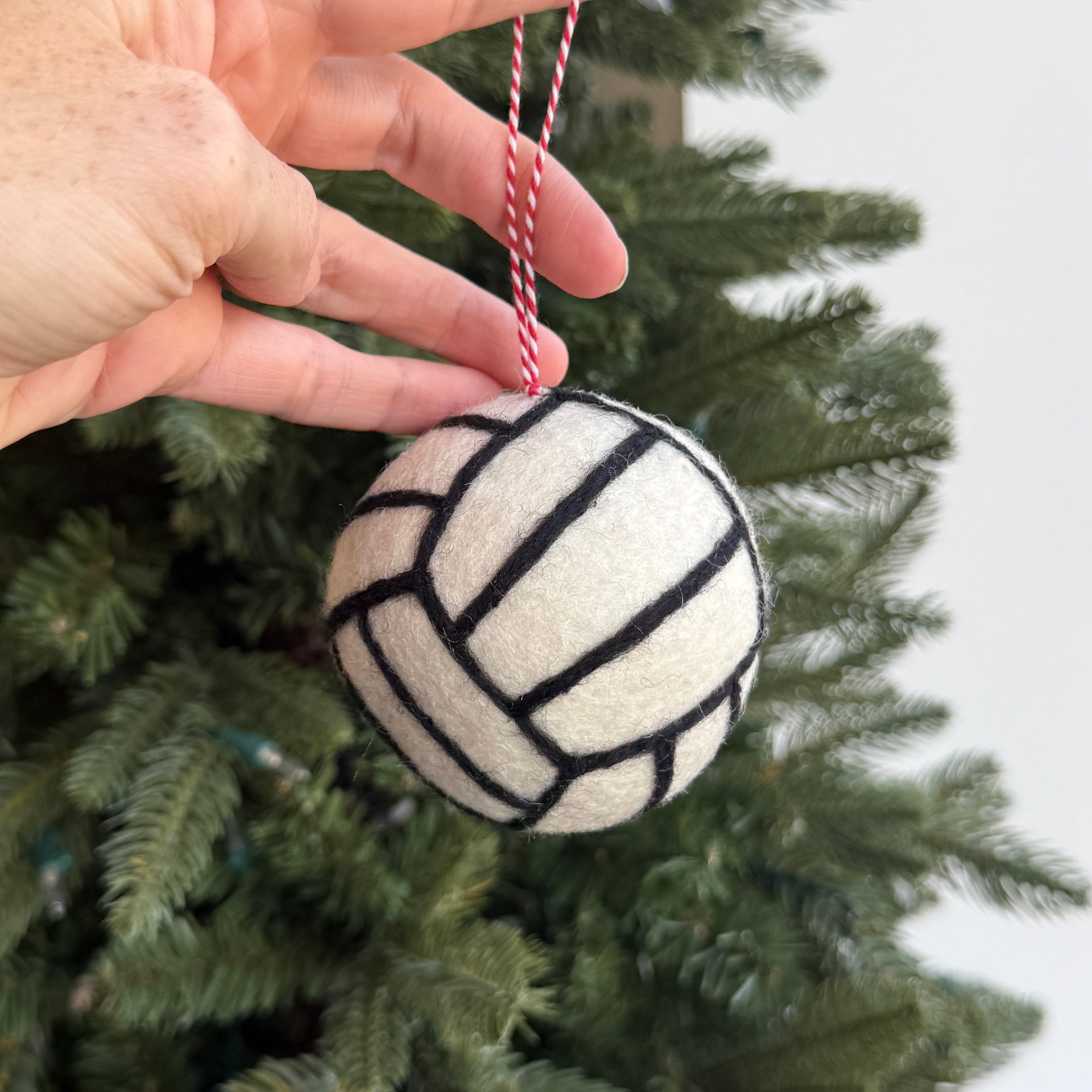 A hand holds a felt Christmas ornament shaped like a volleyball, featuring black stitched lines on an off-white felt surface, hanging from a red and white striped string in front of a green Christmas tree.