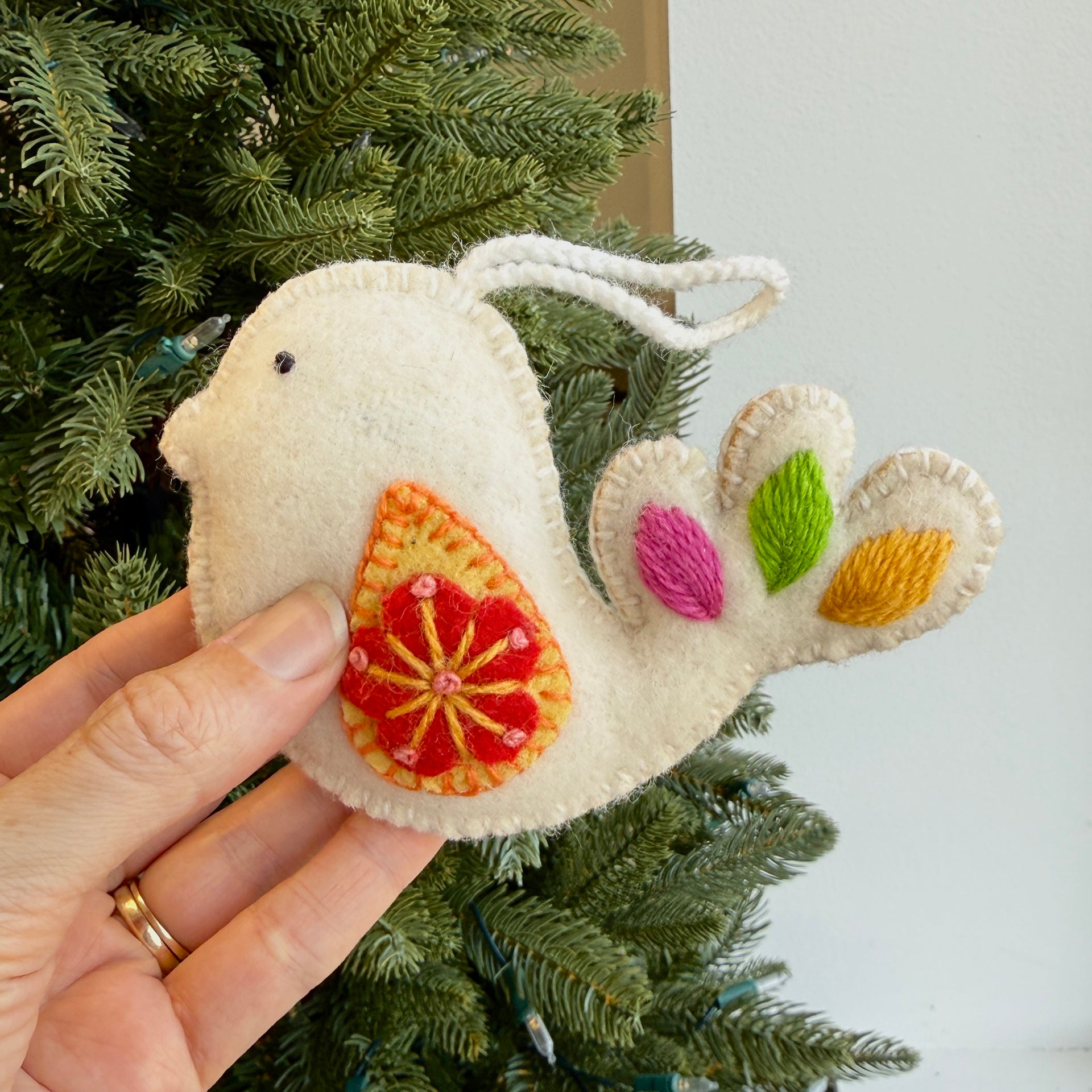 Hand holding a decorative bird ornament with colorful details against a Christmas tree background.