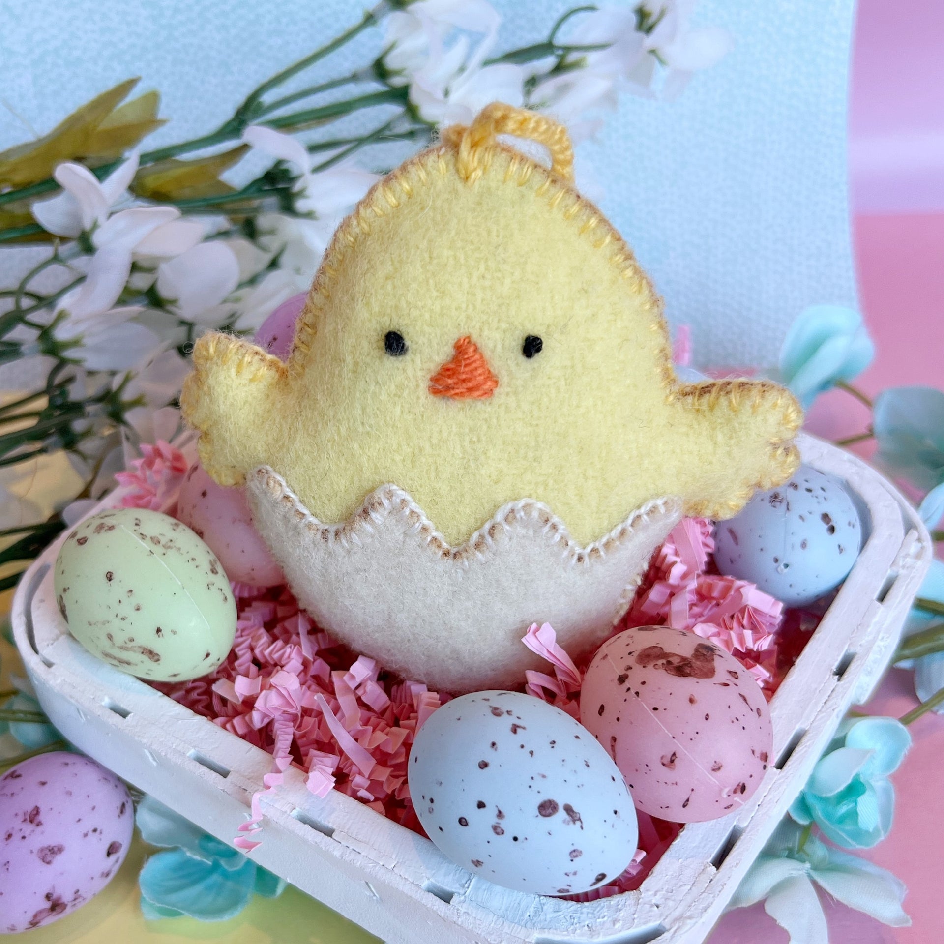Handmade baby chick Easter ornament in basket surrounded by pastel eggs.