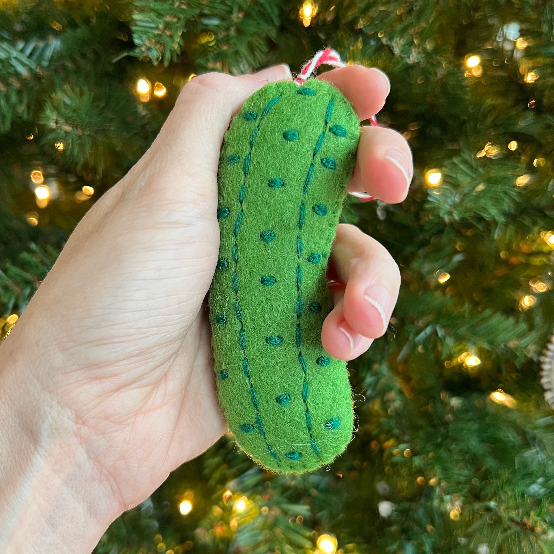 Holding a felt pickle Ornaments 4 Orphans Christmas ornament in front of the tree